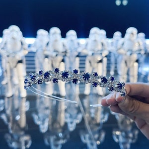 Puede incluir: Una diadema plateada con pedrería azul y transparente. La diadema está decorada con un patrón repetitivo de pequeñas flores.