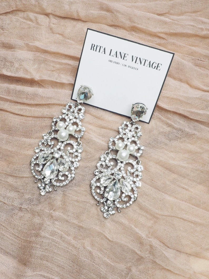 May include: A pair of silver dangle earrings with a crystal and pearl design. The earrings are on a pink fabric background. The card in the image reads "Rita Lane Vintage, Orlando, Los Angeles."