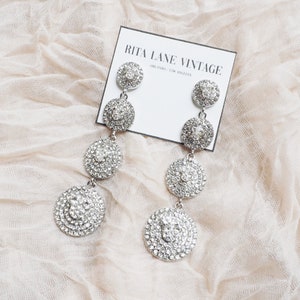 May include: A pair of silver dangle earrings with multiple layers of round, clear rhinestones. The earrings are displayed on a white card with the text "RITA LANE VINTAGE ORLANDO / LOS ANGELES".