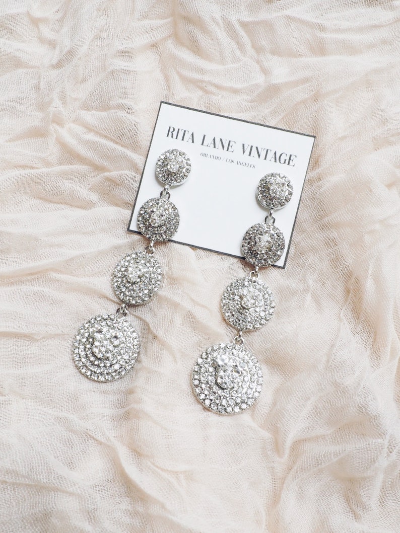 May include: A pair of silver dangle earrings with multiple layers of round, clear rhinestones. The earrings are displayed on a white card with the text "RITA LANE VINTAGE ORLANDO / LOS ANGELES".