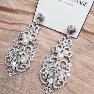 May include: A pair of silver dangle earrings with clear crystal and pearl accents. The earrings are on a pink fabric background. The text "RITA LANE VINTAGE" is visible on a white card.