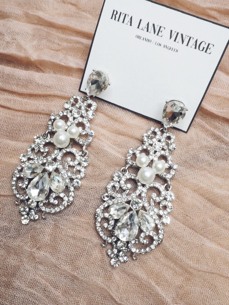 May include: A pair of silver dangle earrings with clear crystal and pearl accents. The earrings are on a pink fabric background. The text "RITA LANE VINTAGE" is visible on a white card.