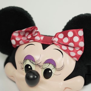 Pu&ograve; includere: Una testa di peluche di Minnie Mouse con orecchie nere, un fiocco rosso con pois bianchi e un viso rosa con ombretto e ciglia viola.