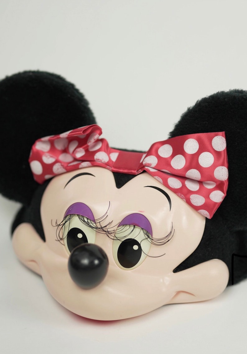 Pu&ograve; includere: Una testa di peluche di Minnie Mouse con orecchie nere, un fiocco rosso con pois bianchi e un viso rosa con ombretto e ciglia viola.