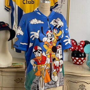 May include: A blue cycling jersey with a white zipper and a colorful print of Mickey Mouse, Donald Duck, Goofy, and Pluto riding bicycles. The jersey has the text "Mickey's Bicycle Club" and "Giordana" printed on it.