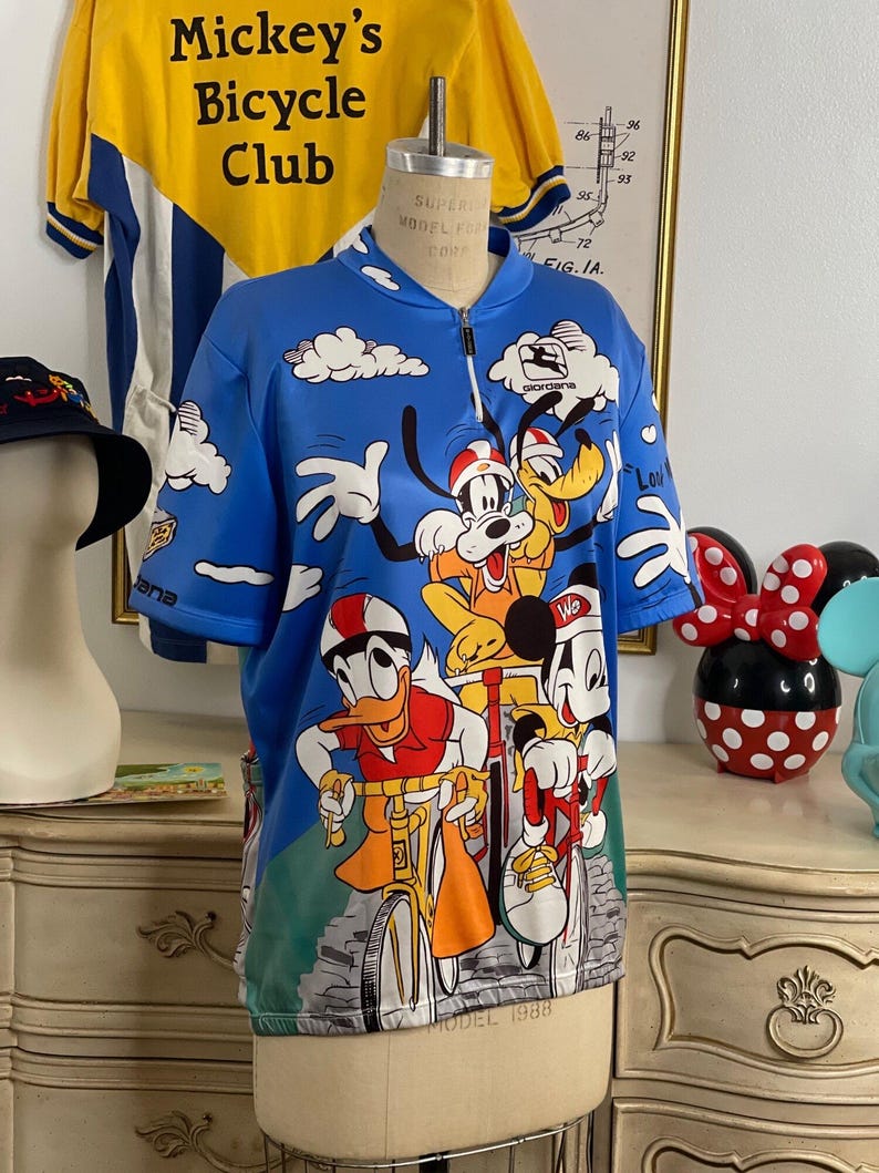 May include: A blue cycling jersey with a white zipper and a colorful print of Mickey Mouse, Donald Duck, Goofy, and Pluto riding bicycles. The jersey has the text "Mickey's Bicycle Club" and "Giordana" printed on it.