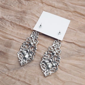May include: A pair of silver dangle earrings with intricate filigree designs and clear crystal accents. The earrings are displayed on a white card with the earring backs visible.
