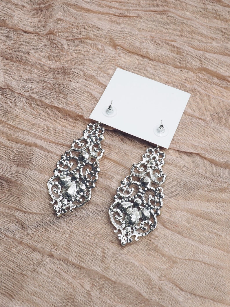 May include: A pair of silver dangle earrings with intricate filigree designs and clear crystal accents. The earrings are displayed on a white card with the earring backs visible.