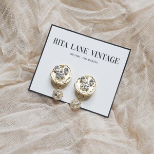 May include: A pair of gold-tone earrings with a round, textured design and clear rhinestones. The earrings are hanging from a small, clear crystal bead.