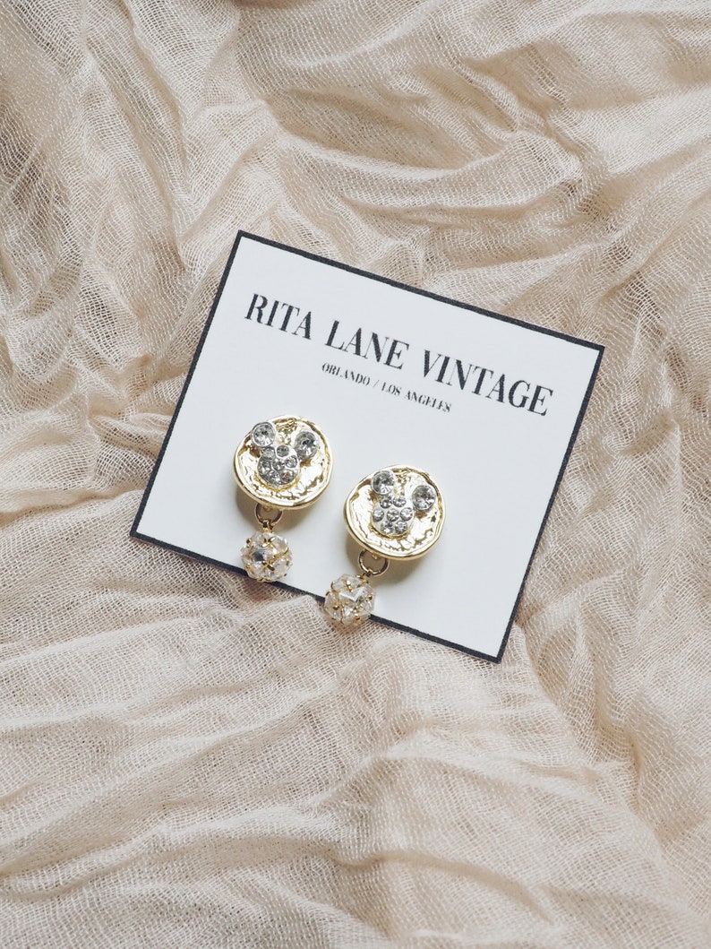 May include: A pair of gold-tone earrings with a round, textured design and clear rhinestones. The earrings are hanging from a small, clear crystal bead.