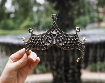 May include: A black jeweled crown with a gold base, held in a hand against a blurred background of a fountain.