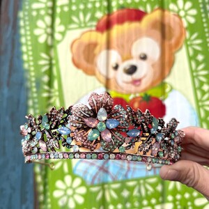 May include: A delicate crown with a floral design featuring pink, blue, and green rhinestones. The crown is made of metal and has a gold finish.