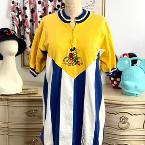 May include: Yellow, blue, and white striped short-sleeved shirt with a yellow collar and a zipper. The shirt features a Mickey Mouse graphic on the front with Mickey riding a bicycle.