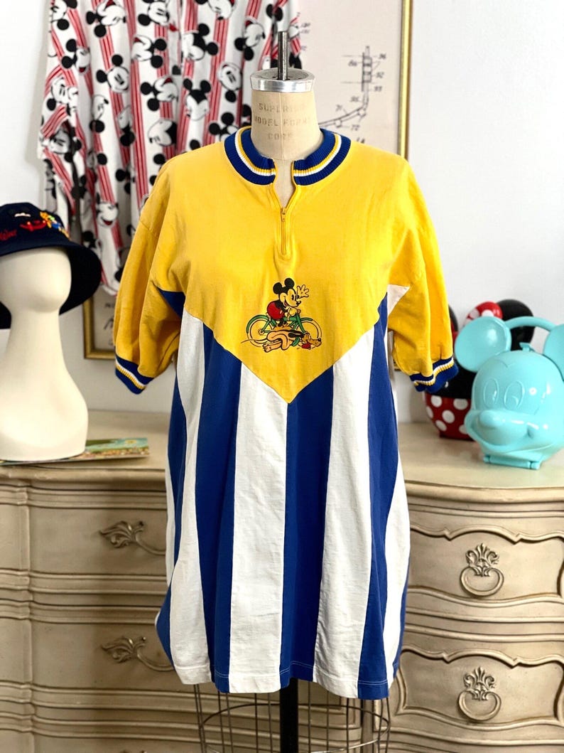 May include: Yellow, blue, and white striped short-sleeved shirt with a yellow collar and a zipper. The shirt features a Mickey Mouse graphic on the front with Mickey riding a bicycle.