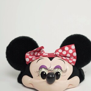 Pu&ograve; includere: Una testa di Minnie Mouse in peluche nera e bianca con un fiocco rosso con pois bianchi. La testa ha un viso rosa con occhi neri, un naso nero e sopracciglia viola.