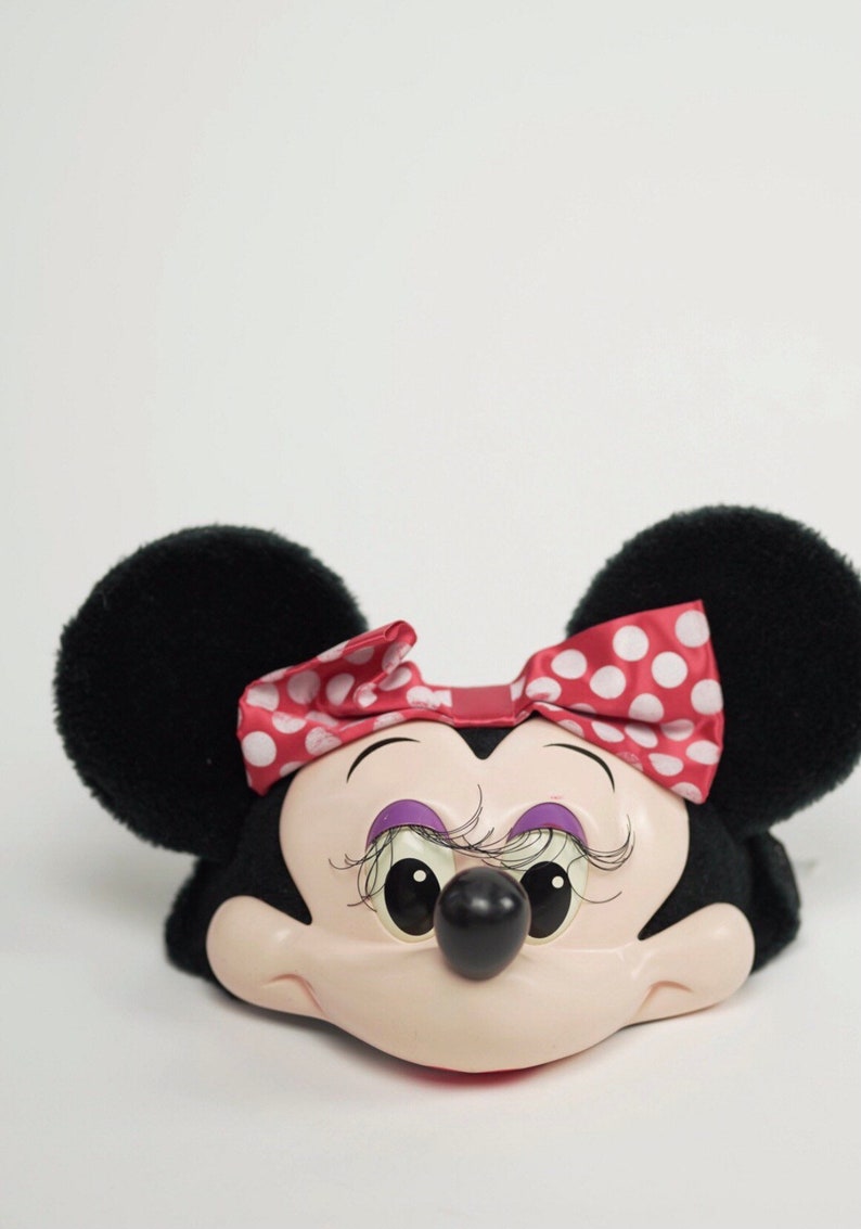 Pu&ograve; includere: Una testa di Minnie Mouse in peluche nera e bianca con un fiocco rosso con pois bianchi. La testa ha un viso rosa con occhi neri, un naso nero e sopracciglia viola.