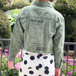 May include: A green denim jacket with a vintage Disneyland "Date Nite" advertisement printed on the back. The advertisement features a cartoon character in a teacup.