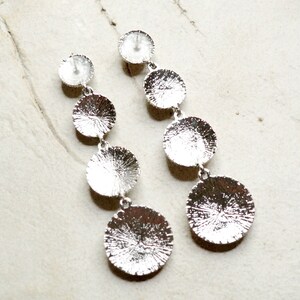 May include: Silver dangle earrings with four round discs of varying sizes. The discs have a textured surface.
