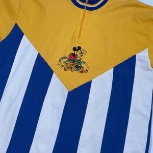 May include: A yellow and blue striped vintage cycling jersey with a Mickey Mouse graphic riding a bicycle. The jersey has a zipper closure and a white collar with blue trim.
