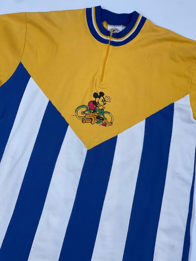May include: A yellow and blue striped vintage cycling jersey with a Mickey Mouse graphic riding a bicycle. The jersey has a zipper closure and a white collar with blue trim.