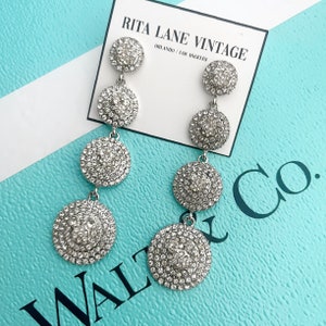 May include: A pair of silver dangle earrings with four round crystal-encrusted charms. The earrings are displayed on a turquoise box with the text "Rita Lane Vintage Orlando / Los Angeles" and "Walt & Co."