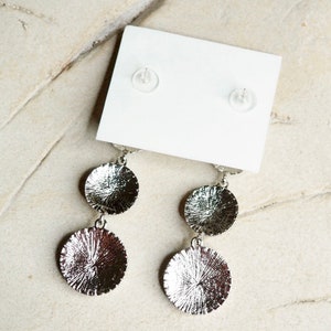 May include: A pair of silver dangle earrings with two round discs on each earring. The discs have a textured surface.