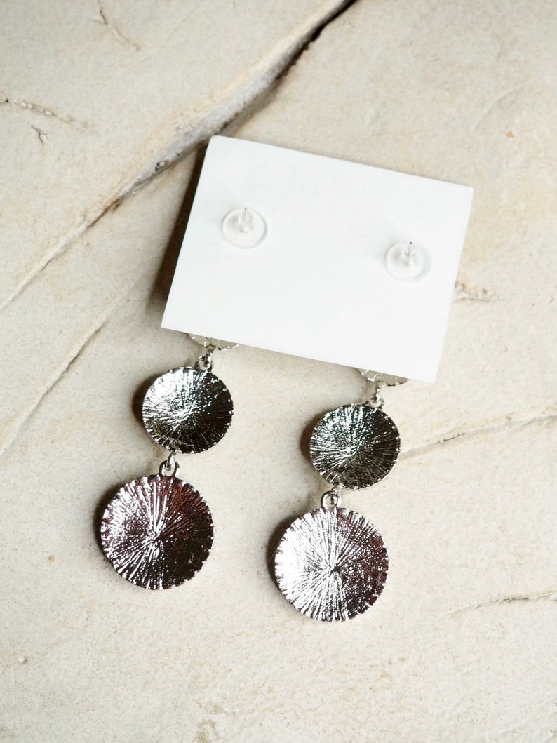 May include: A pair of silver dangle earrings with two round discs on each earring. The discs have a textured surface.
