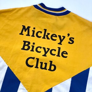 May include: Yellow, white, and blue striped shirt with a yellow panel on the back. The panel has the text "Mickey's Bicycle Club" in black.