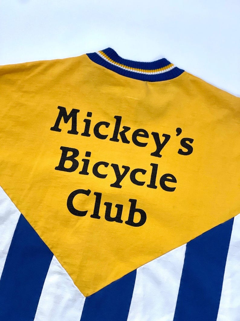 May include: Yellow, white, and blue striped shirt with a yellow panel on the back. The panel has the text "Mickey's Bicycle Club" in black.