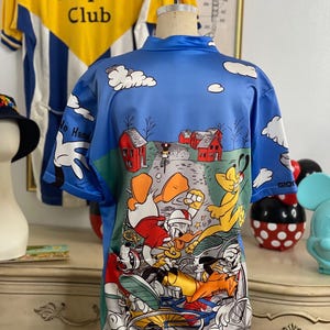 May include: A blue cycling jersey with a cartoon print of Mickey Mouse, Donald Duck, Goofy, and Pluto riding bicycles. The jersey has a white collar and short sleeves. The print features a blue sky with white clouds and red houses in the background.