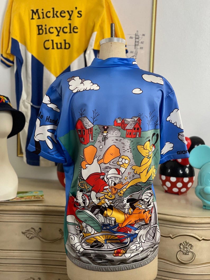 May include: A blue cycling jersey with a cartoon print of Mickey Mouse, Donald Duck, Goofy, and Pluto riding bicycles. The jersey has a white collar and short sleeves. The print features a blue sky with white clouds and red houses in the background.