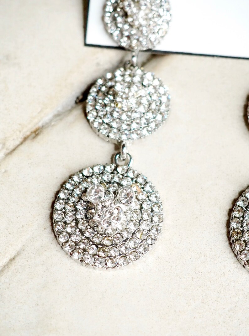 May include: Silver dangle earrings with three round crystal-encrusted charms. The bottom charm features a Mickey Mouse silhouette.