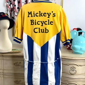 May include: A yellow and blue striped cycling jersey with a yellow panel on the back. The panel has the text "Mickey's Bicycle Club" in black.