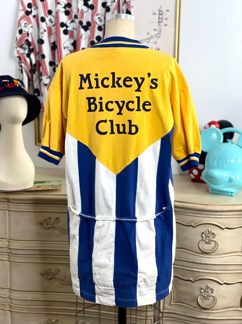May include: A yellow and blue striped cycling jersey with a yellow panel on the back. The panel has the text "Mickey's Bicycle Club" in black.