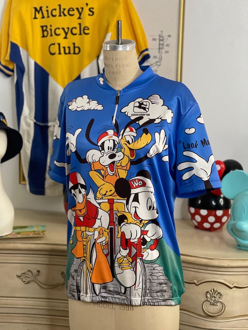 May include: A blue cycling jersey with a print of Mickey Mouse, Donald Duck, Goofy, and Pluto riding bicycles. The jersey has a zipper and the text "Mickey's Bicycle Club" and "Look Ma, No Hands!" printed on it.