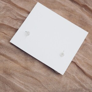 May include: A pair of small, clear crystal earrings on a white card, set against a brown burlap background.