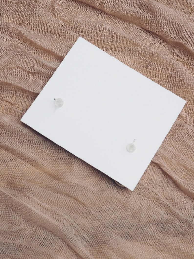 May include: A pair of small, clear crystal earrings on a white card, set against a brown burlap background.