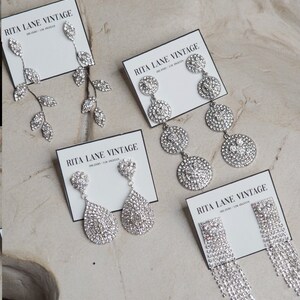 May include: Four pairs of silver rhinestone earrings on white cards with the text "RITA LANE VINTAGE" printed on them. The earrings are all different styles, including teardrop, leaf, and circle designs.