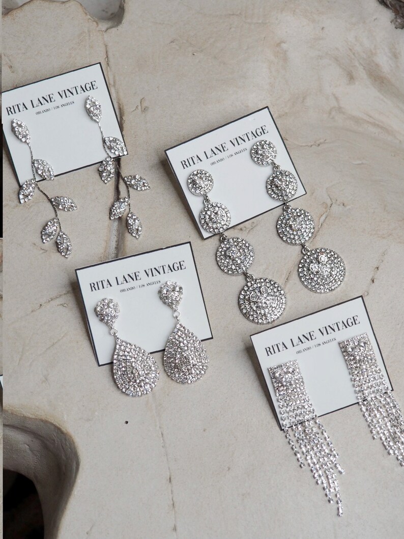 May include: Four pairs of silver rhinestone earrings on white cards with the text "RITA LANE VINTAGE" printed on them. The earrings are all different styles, including teardrop, leaf, and circle designs.