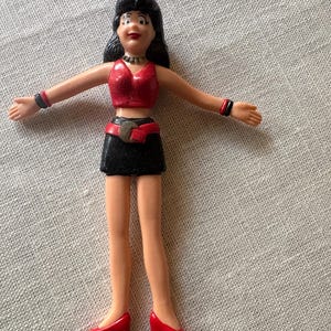 May include: A plastic figurine of a person with black hair, wearing a red glittery crop top, a black skirt with a red belt, and red high-heeled shoes. The figure has black bracelets on both wrists and a silver-coloured necklace around the neck.