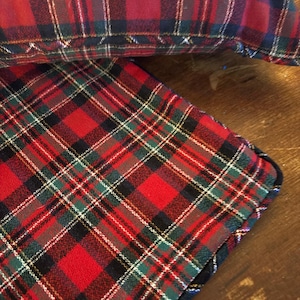 May include: Close-up of two plaid fabric items. The pattern features a red base with black, green, and white intersecting lines, creating a classic tartan design. One item appears to be a pillow, the other a flat piece of fabric.
