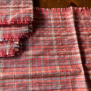May include: A set of rectangular fabric placemats featuring a red, brown, and blue plaid pattern. Each placemat has a textured weave and frayed edges along the sides, creating a soft, unfinished look across the entire collection of linens.