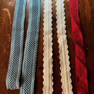 May include: Five strips of decorative trim arranged vertically. Two strips are blue with a diamond-patterned weave, two strips are cream-colored with a central braided texture and scalloped edges, and one strip is solid red with a fine woven texture.