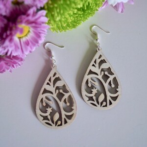 May include: A pair of teardrop-shaped wooden earrings with a delicate floral design. The earrings are a light beige colour and have silver hooks.