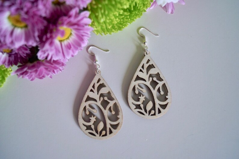May include: A pair of teardrop-shaped wooden earrings with a delicate floral design. The earrings are a light beige colour and have silver hooks.