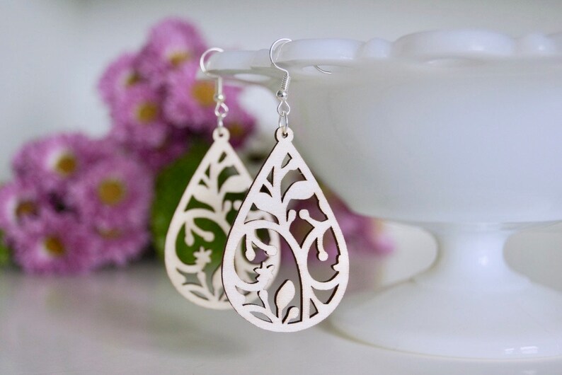 May include: A pair of wooden earrings with a teardrop shape and intricate floral cutouts. The earrings are made of light-coloured wood and have silver hooks.