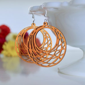 May include: A pair of large, round, wooden earrings with a geometric, lattice-like design. The earrings are a light brown color and have silver hooks.
