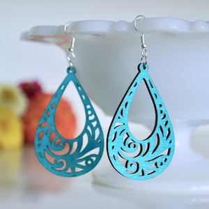May include: Teal blue dangle earrings with a cutout floral design. The earrings are made of wood and have silver hooks.