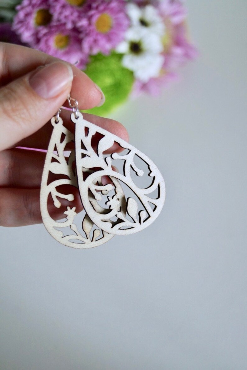 May include: A pair of wooden earrings shaped like teardrops with a delicate floral cut-out design. The earrings are a light natural wood colour.