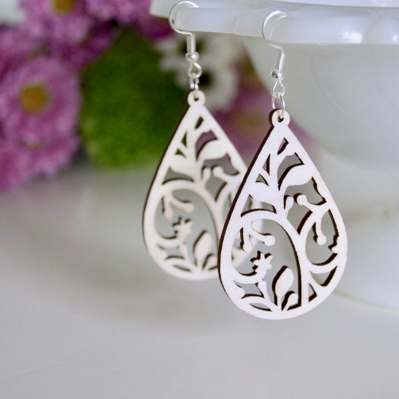 May include: A pair of teardrop-shaped wooden earrings with a delicate floral cutout design. The earrings are white and have a natural wood finish.